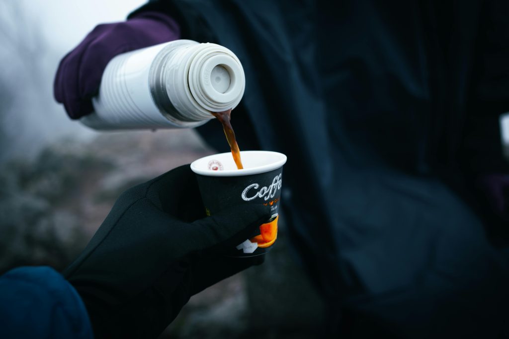 A person pours coffee into a cup from a thermos in a cool outdoor setting, creating a cozy atmosphere.